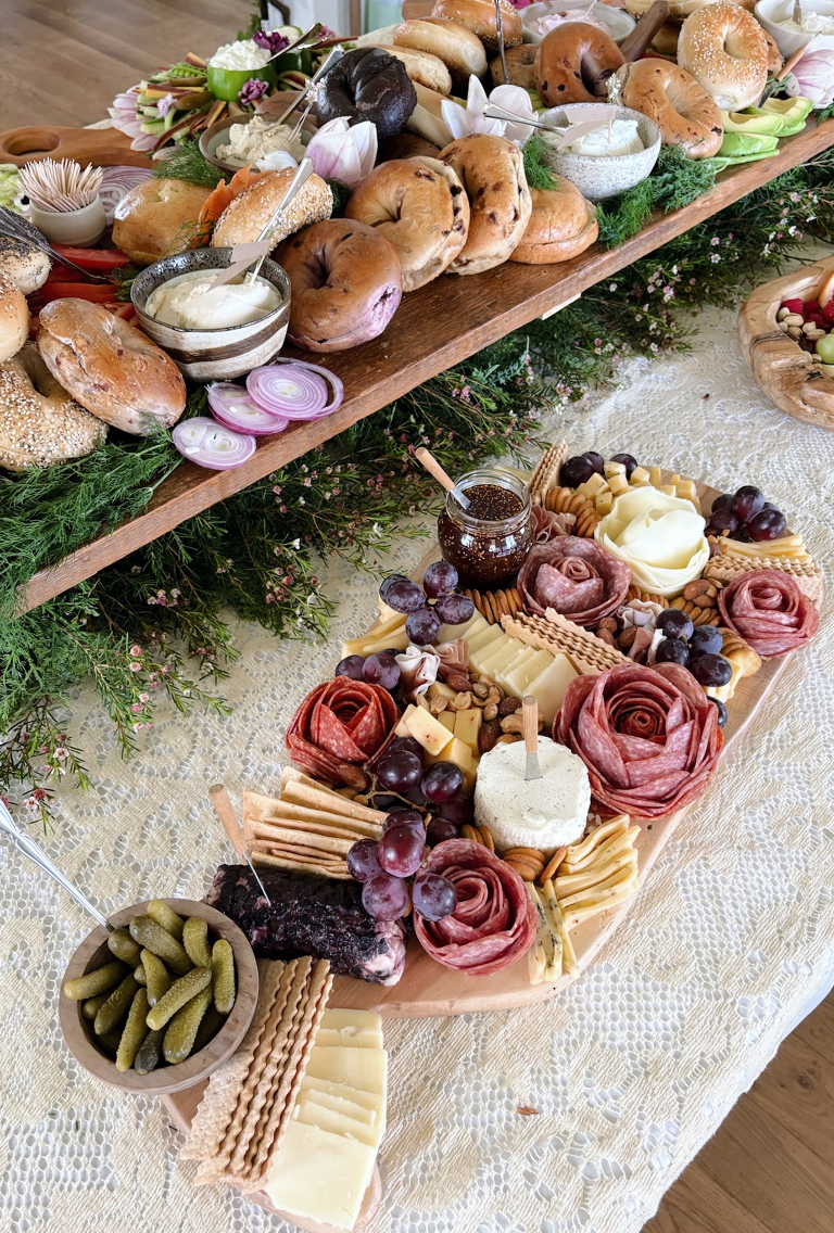 table with bagel platter and charcuterie board on it with greenery on table
