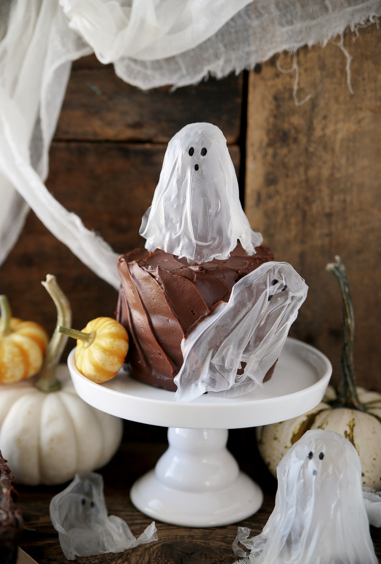 chocolate cake on white cake stand with ghost decorations on cake and mini pumpkins around