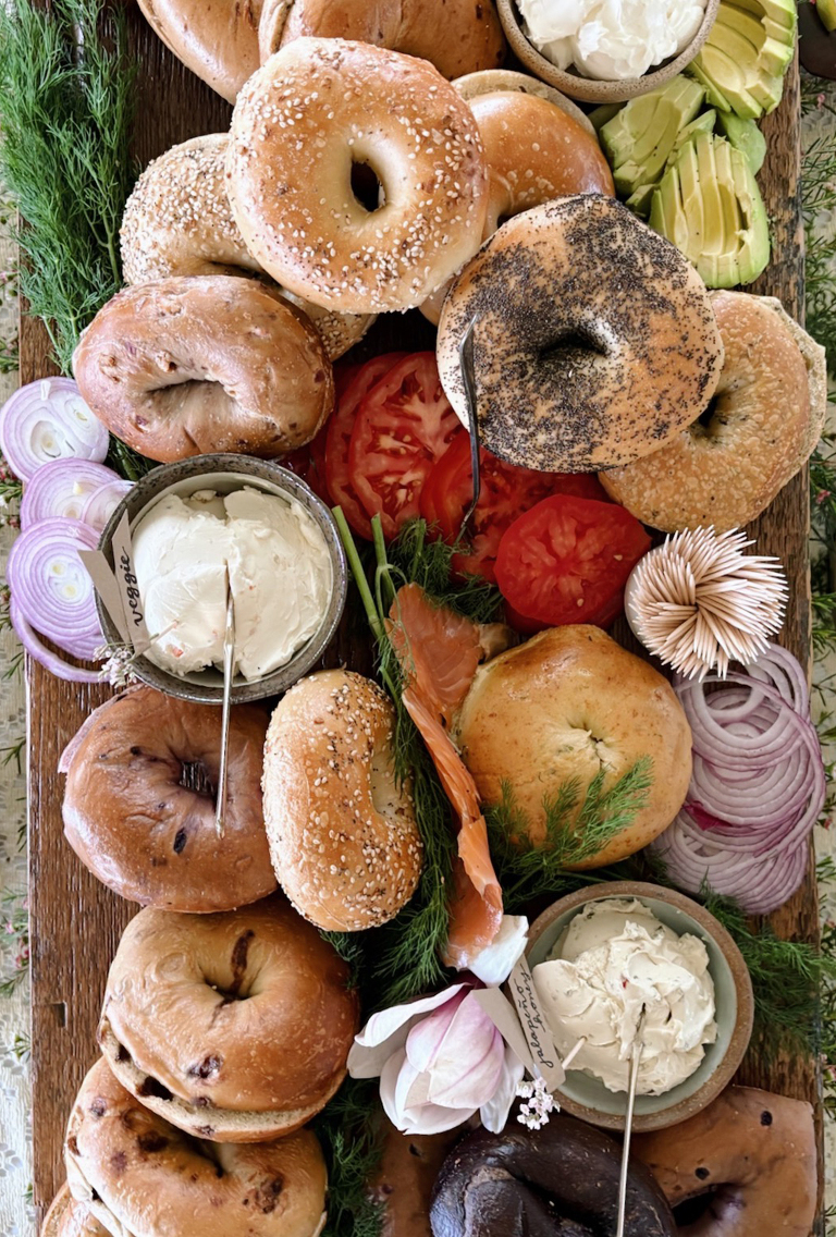 bagel board with bagels, tomatoes, dill, avocados, onions, lox and bowls of cream cheese on board