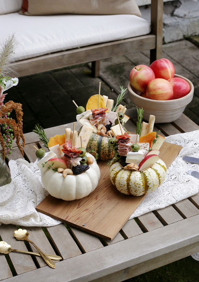 three individual charcuterie boards arranged inside carved out mini pumpkins on wooden board next to bowl of apples