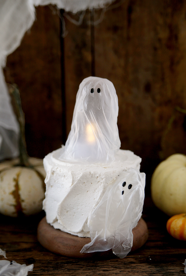 ghost decoration on small white cake with another ghost on side of cake with mini pumpkins in background