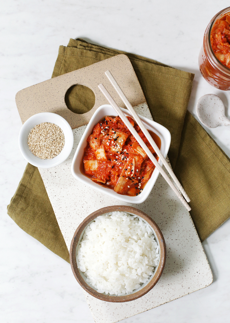 kimchi in white square bowl next to bowl of rice on ceramic cutting board