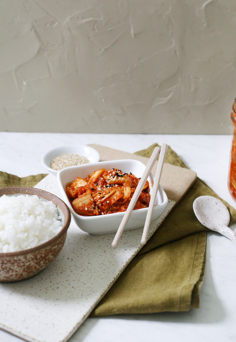 bowl of kimchi with chopsticks rested on the edge on a ceramic cutting board on top of green linen kitchen towel