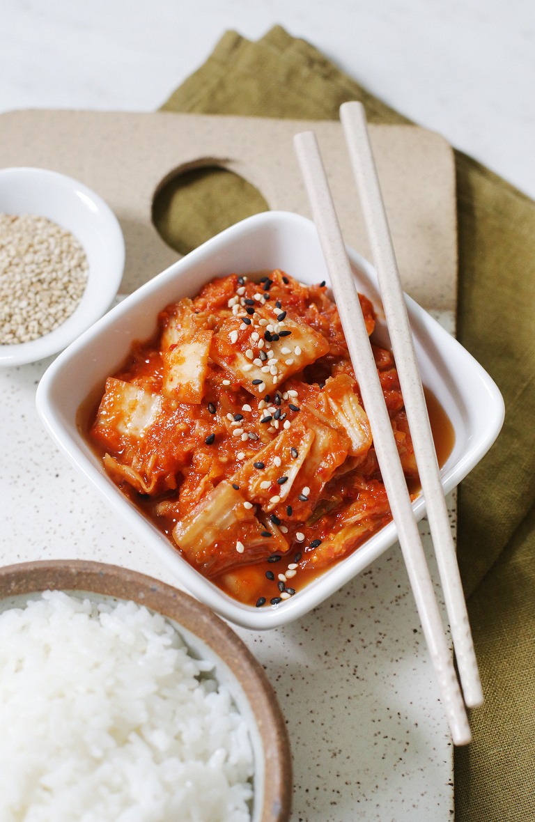 bowl of kimchi with chopsticks next to bowl of rice and sesame seeds on ceramic cutting board