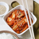 bowl of kimchi with chopsticks next to bowl of rice and sesame seeds on ceramic cutting board