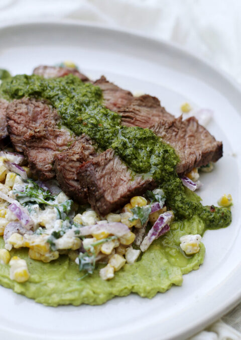 sliced steak over corn and onion salad with mashed avocado on white plate