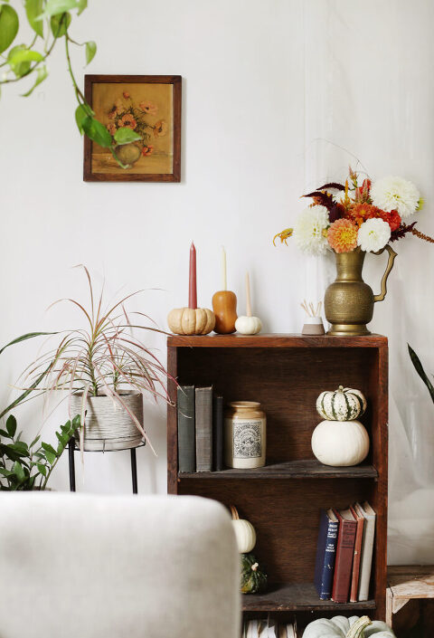 brown shelf decorated with pumpkins, flowers and books in front of white wall with small floral art
