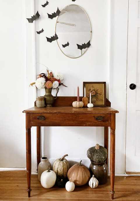 pumpkins and flowers surrounding table in front of white wall with oval mirror and black cut out bats on the wall