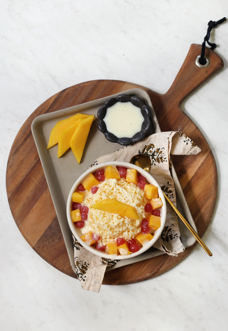 bowl of shave mango ice with fresh mango and boba in bowl on tray next to ingredients on top of round wooden cutting board