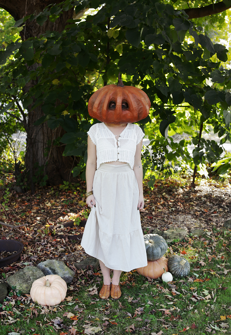 girl in white outfit in pumpkin mask standing next to pumpkins in front of trees