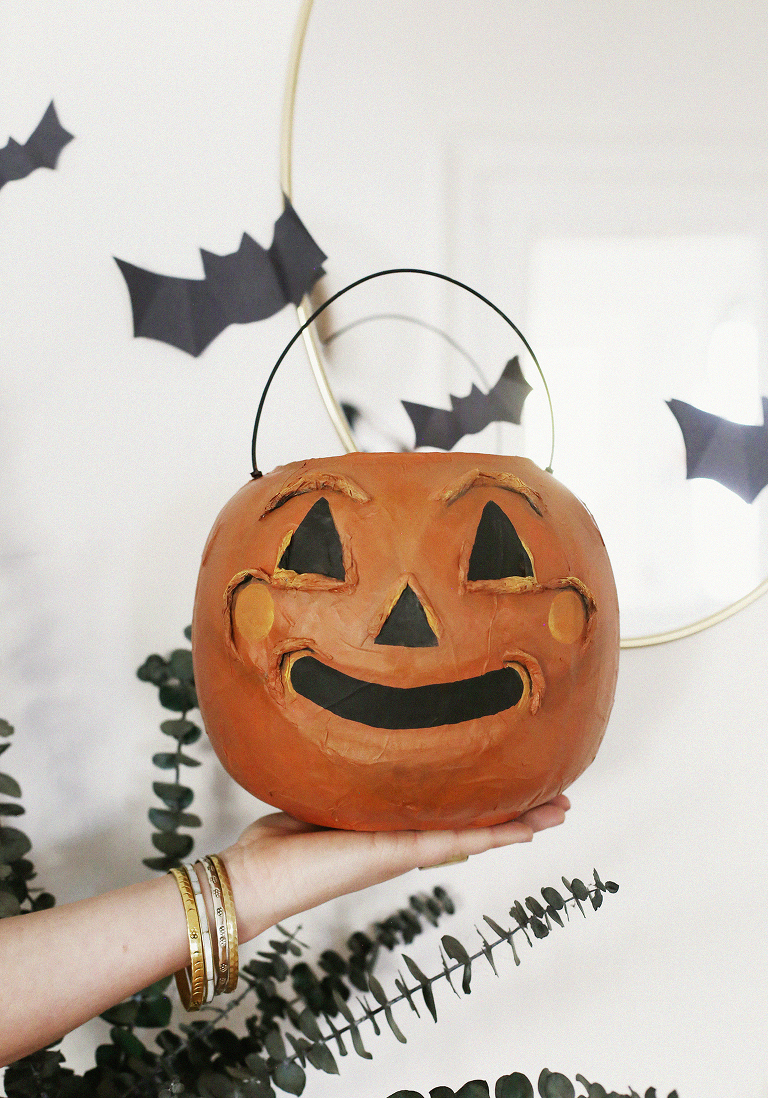 pumpkin treat bucket held in a hand in front of white wall with cut out black bats