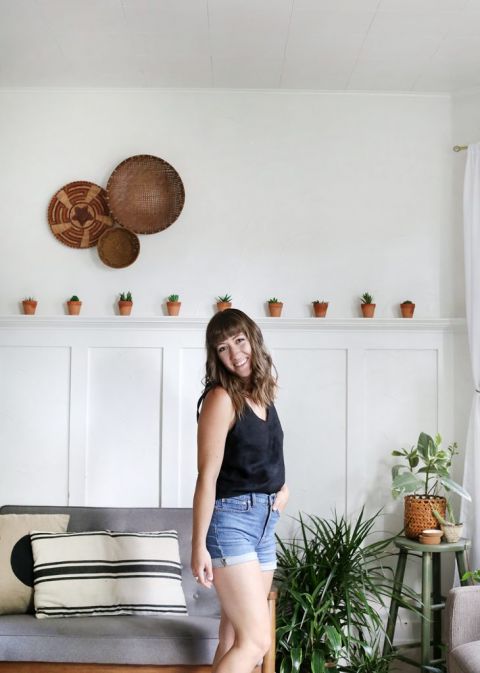 woman in black shirt and jean shorts in front of grey couch and white wall with plants