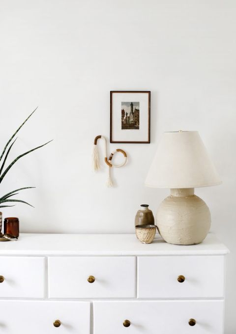 white dresser in white room with cream lamp and ceramics on dresser, fiber art and frame on wall next to lamp