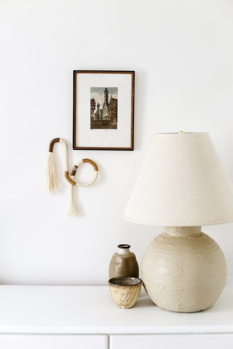 fiber art and frame on white wall next to lamp and neutral ceramics on top of white dresser