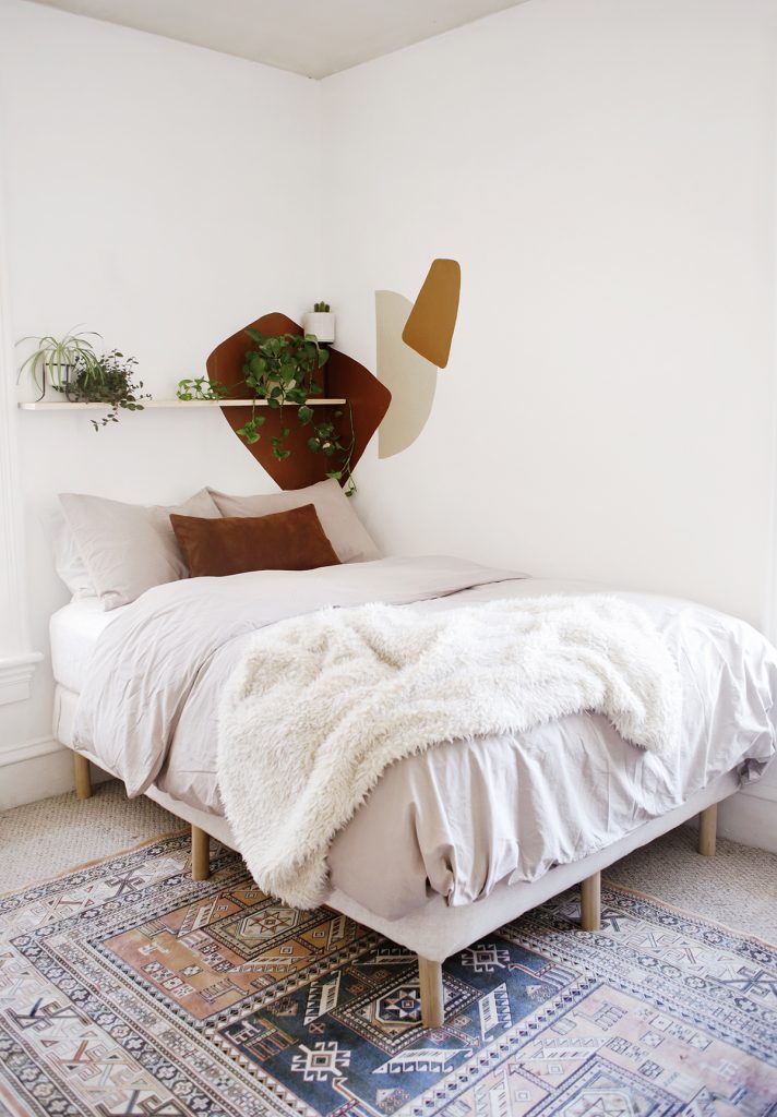 cream bed against white wall with painted abstract shapes and a plant shelf