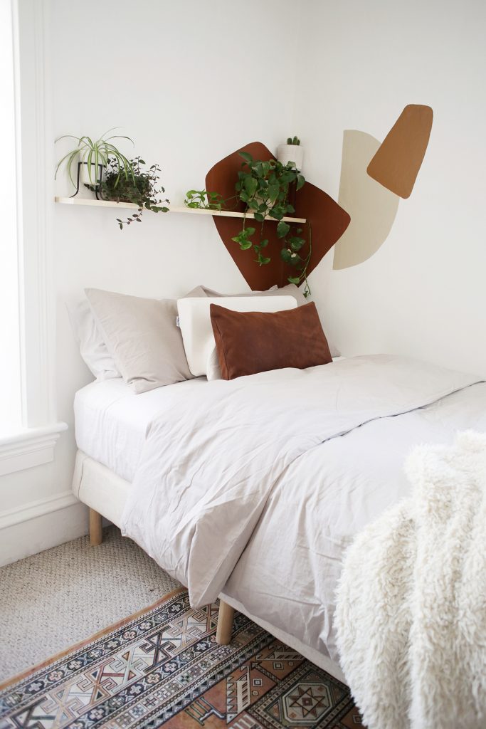 cream bed against white wall with painted abstract shapes and a plant shelf