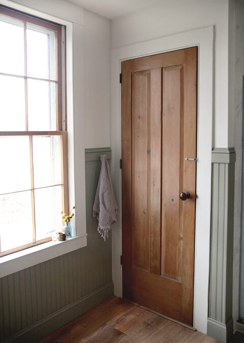 corner of bathroom with wooden door and window with glass bottles sitting on sill