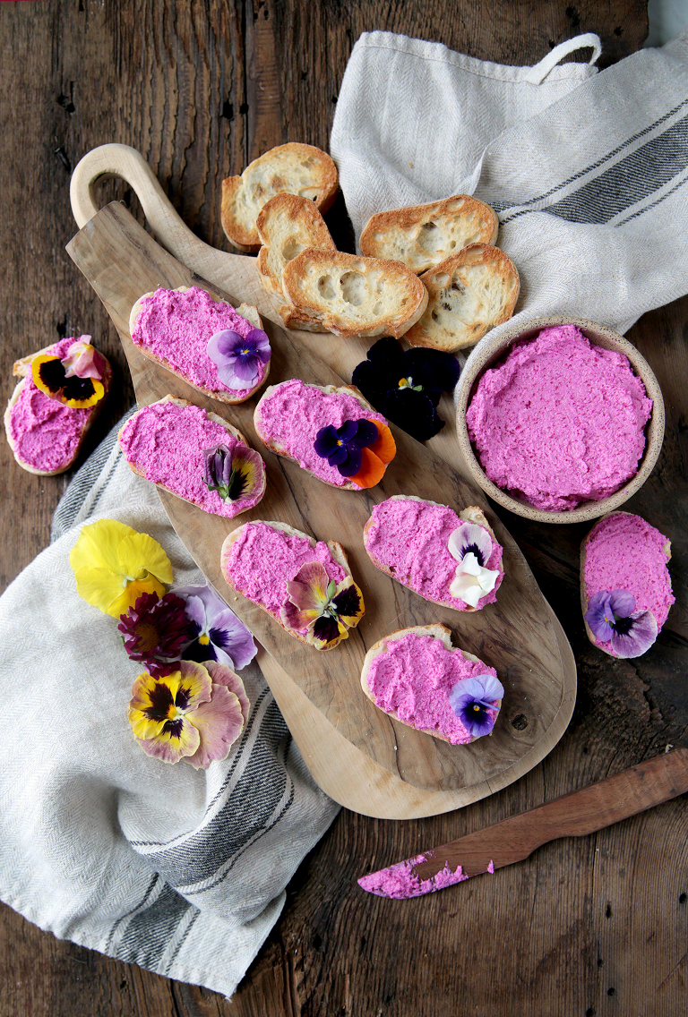 wood cutting board with beet feta goat cheese crostini on it next to bowl of the beet feta goat cheese and plain crostini and edible flowers laying next to cutting board