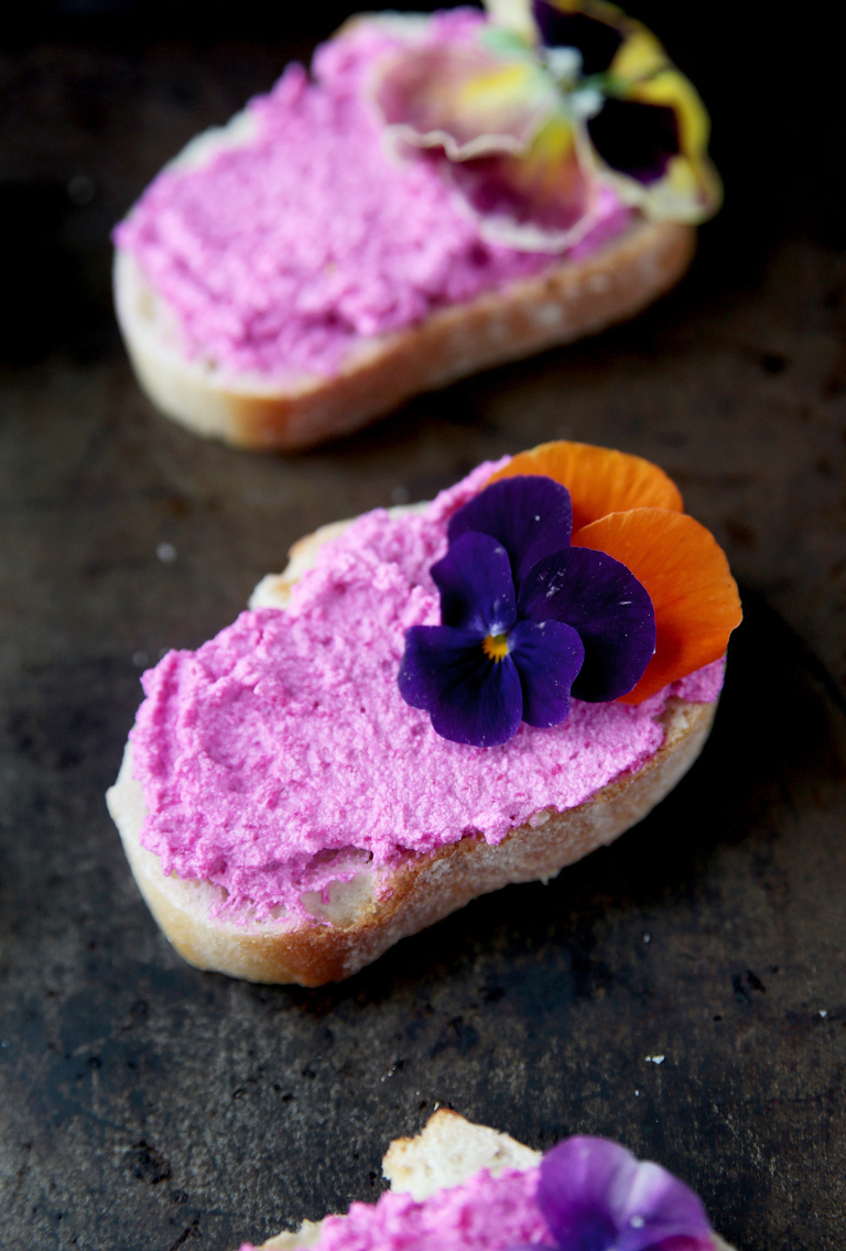 close up photo of beet feta goat cheese crostini with dark purple and orange edible flower on it