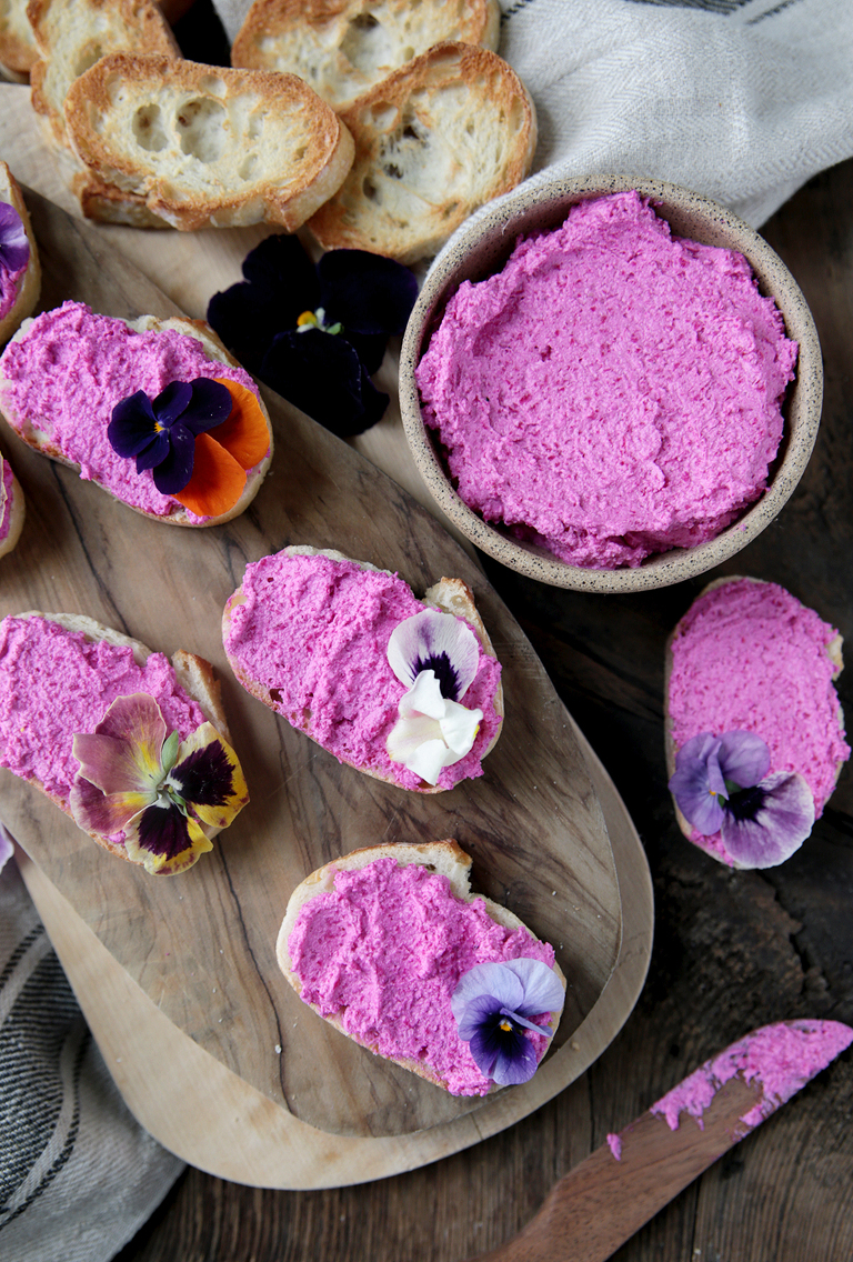 pink beet whipped feta crostini on wood cutting board next to bowl of the beet whipped feta and wood knife laying next to cutting board