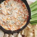skillet filled with buffalo chicken dip with celery chips and bread next to it