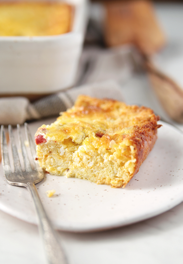 side view of cut piece of cheesy egg casserole on plate with fork next to casserole