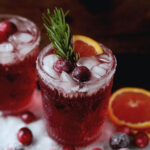 glass of cranberry ginger mocktail with crushed ice in it, sugared cranberries, rosemary stem and small orange wedge. Sugared cranberries and orange slices sit around glass.