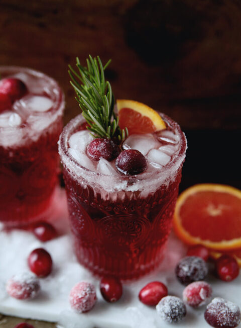glass of cranberry ginger mocktail with crushed ice in it, sugared cranberries, rosemary stem and small orange wedge. Sugared cranberries and orange slices sit around glass.