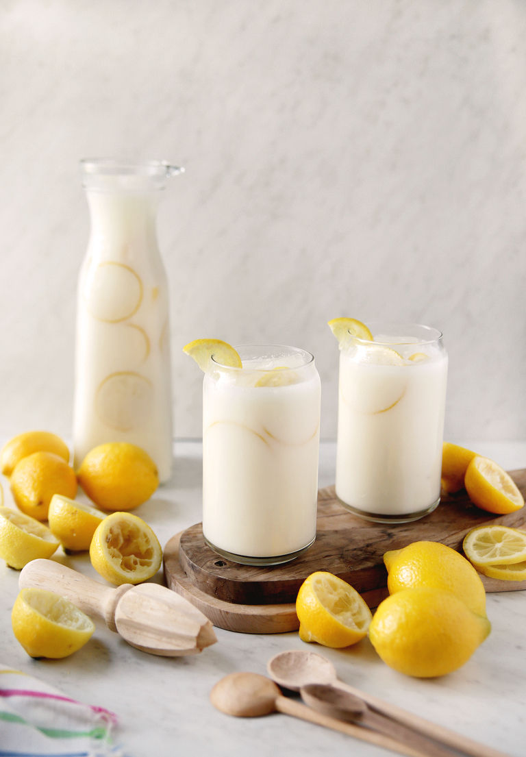 two glasses of creamy lemonade on cutting board next to glass pitcher of lemonade with lemons and squeezed lemons next to it