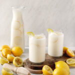 two glasses of creamy lemonade on cutting board next to glass pitcher of lemonade with lemons and squeezed lemons next to it