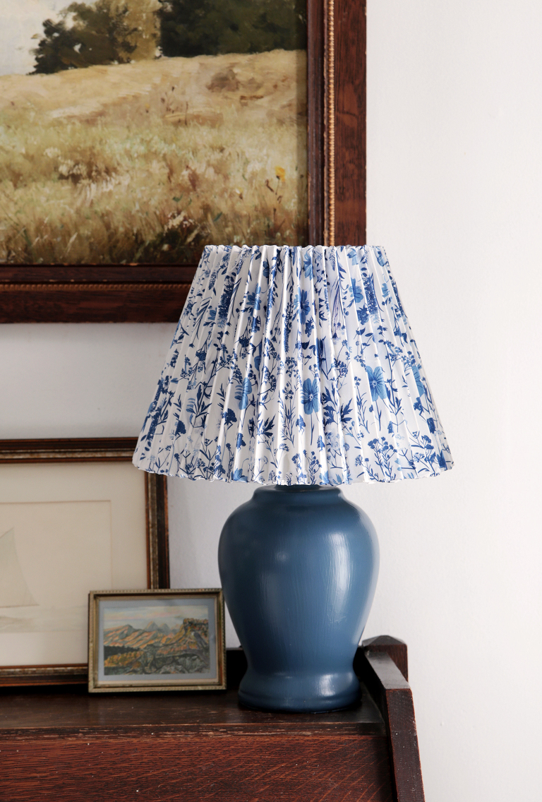 blue floral pleated lampshade on blue lamp next to vintage paintings