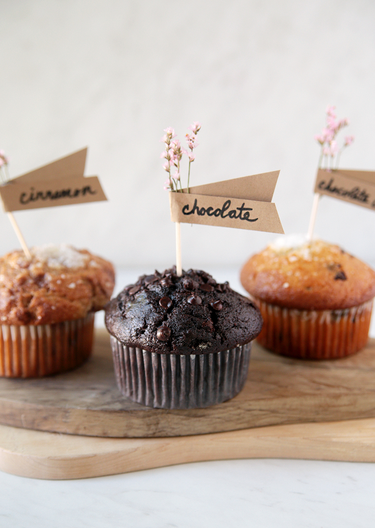 three muffins on wood cutting boards with toothpick food labels stuck in top of muffins