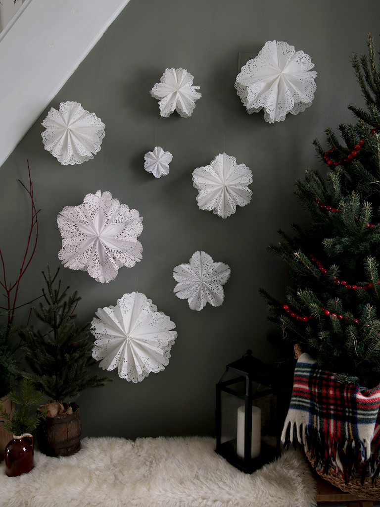8 paper snowflakes hanging on wall next to small christmas tree and lantern on bench