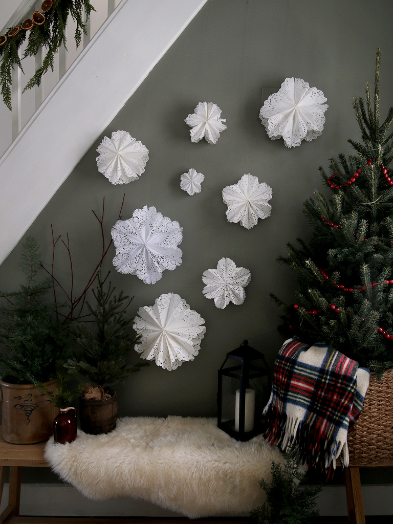 paper doily snowflakes hanging on green wall next to christmas trees on bench with sheepskin fur throw on it