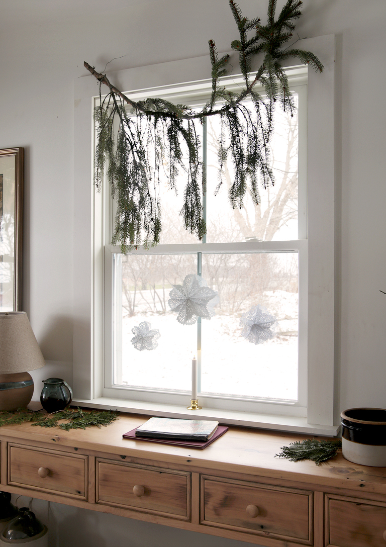 window with paper snowflakes in it with greenery hanging from window and antique wood console table by window