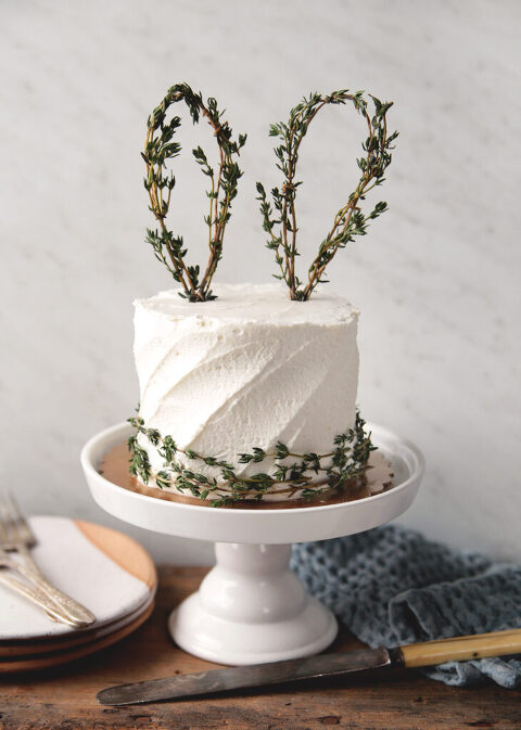 white cake on white cake stand with thyme bunny ears on top of cake with thyme stems wrapped around base of cake