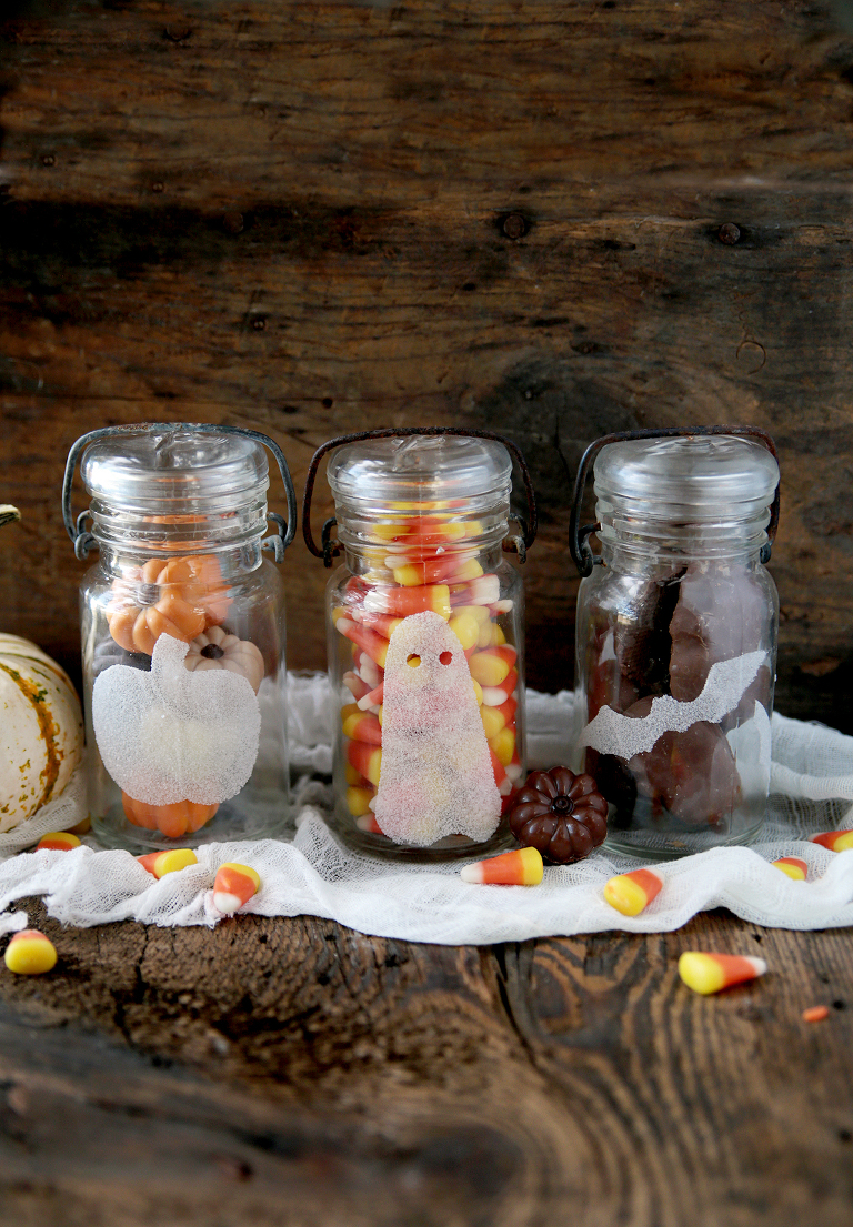 three glass decorative halloween salt jars sitting on cheesecloth on wood board with halloween candy in jars