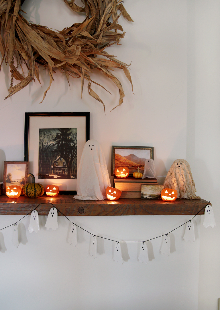 wood mantel decorated for halloween with ghost garland, cheesecloth ghosts, mini pumpkins and orange jack o lanterns with tea lights in them