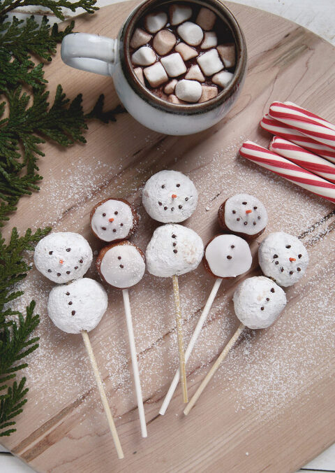 snowman donut holes on sticks laying on wood wearing workbench with hot chocolate and peppermint sticks