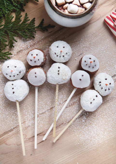 snowman donut holes on sticks on wood wearing boards with powdered sugar dusting on them