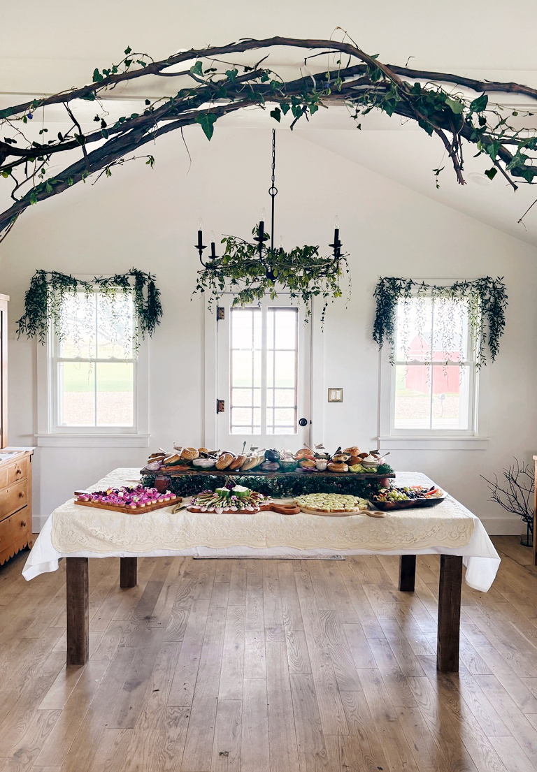 dining room set up for floral bridal shower with greenery hanging from windows and chandelier and bagel bar and food platters on table