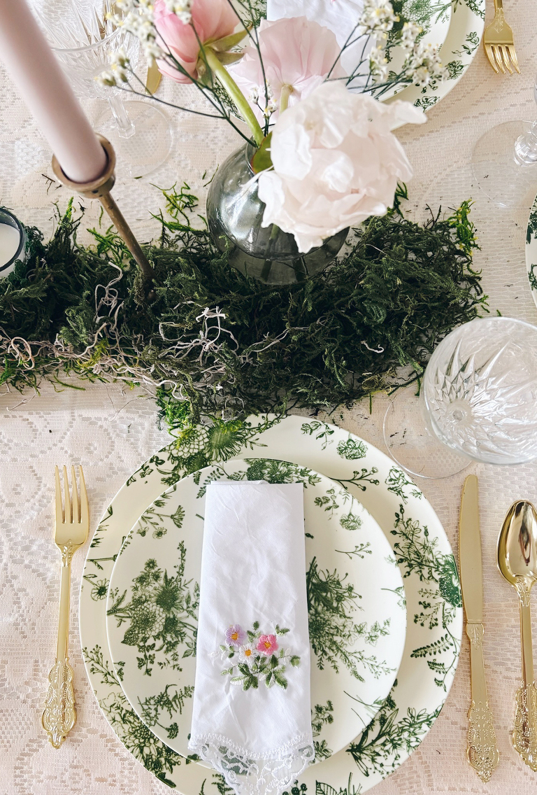 overhead view of place setting with lace table cloth green floral dishes and moss with candles and flowers around it