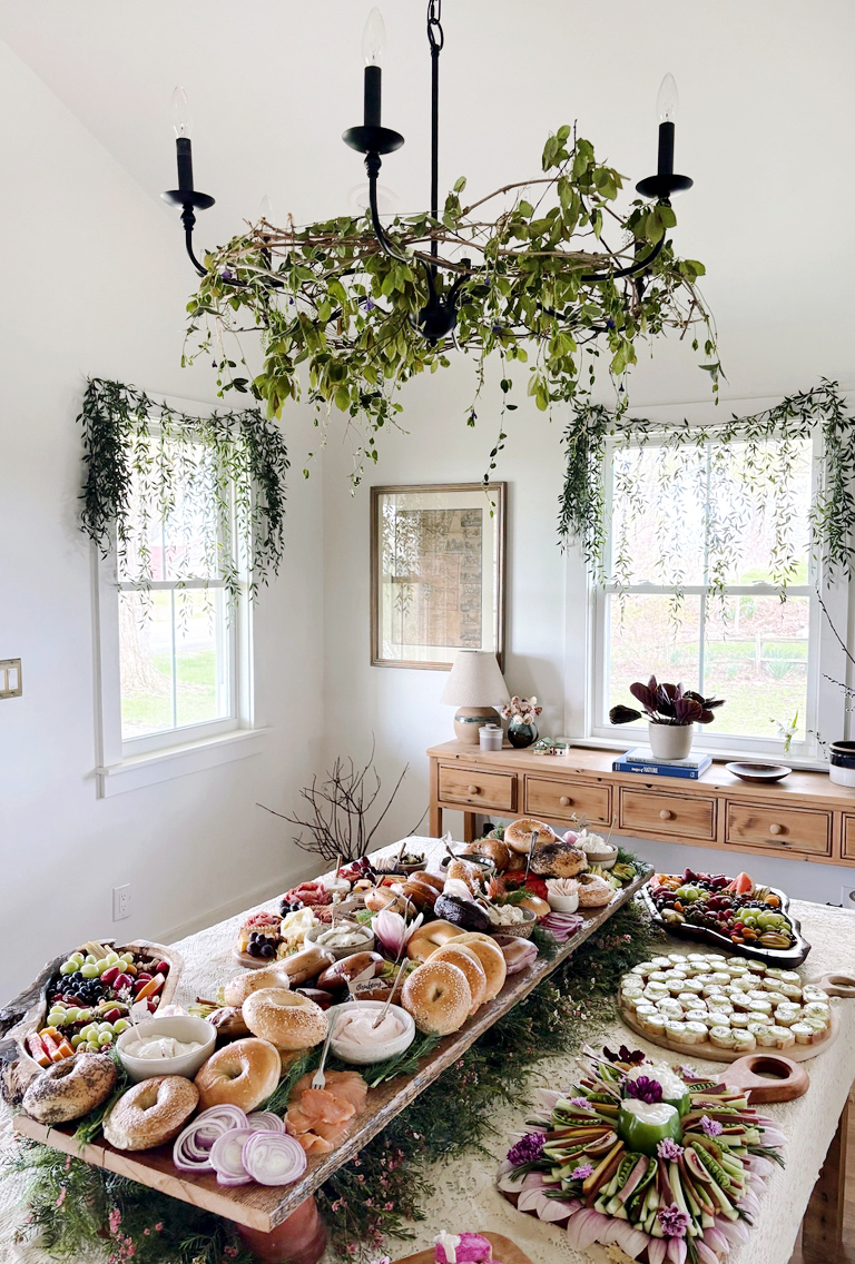 dining room with greenery on windows and chandelier with bagel bar and food platters on table below chandelier