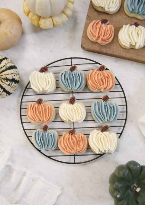 round cooling rack with pumpkin sugar cookies on it frosted in orange, white and blue frosting with small pumpkins next to rack