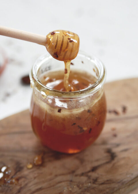 glass jar of honey with red pepper flakes in it with wood honey stick lifting out of it