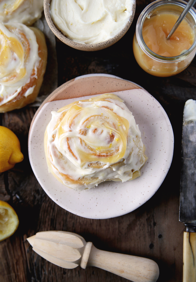lemon sweet roll on ceramic plate topped with lemon frosting and lemon curd with citrus laying next to plate and jar of lemon curd next to plate