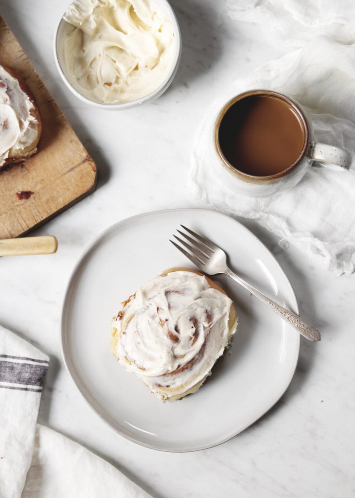 white plate with cinnamon roll and fork on it next to a mug of coffee