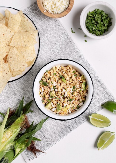 bowl of mexican street corn dip surrounded by tortilla chips, lime wedges, cilantro, bowl of queso fresco and ear of corn
