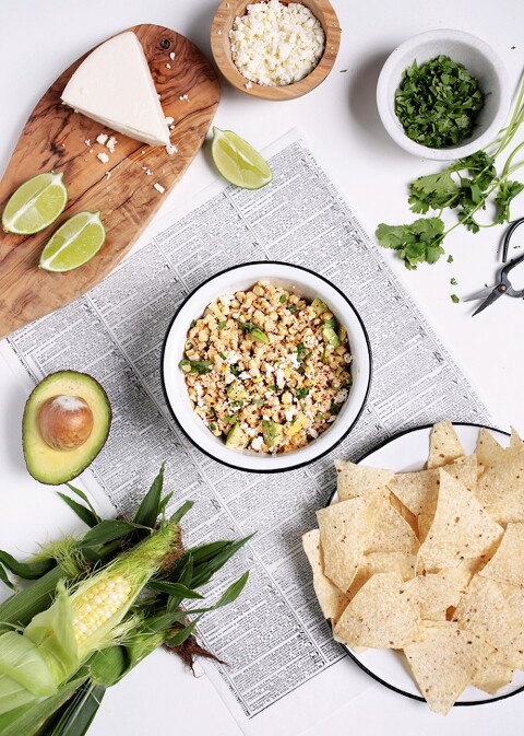 bowl of mexican street corn dip surrounded by tortilla chips, lime wedges, sliced avocado, cilantro and corn on the cob