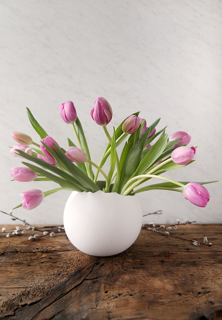 giant plaster easter egg vase with pink tulips in it sitting on wood board with pussy willow branches around it